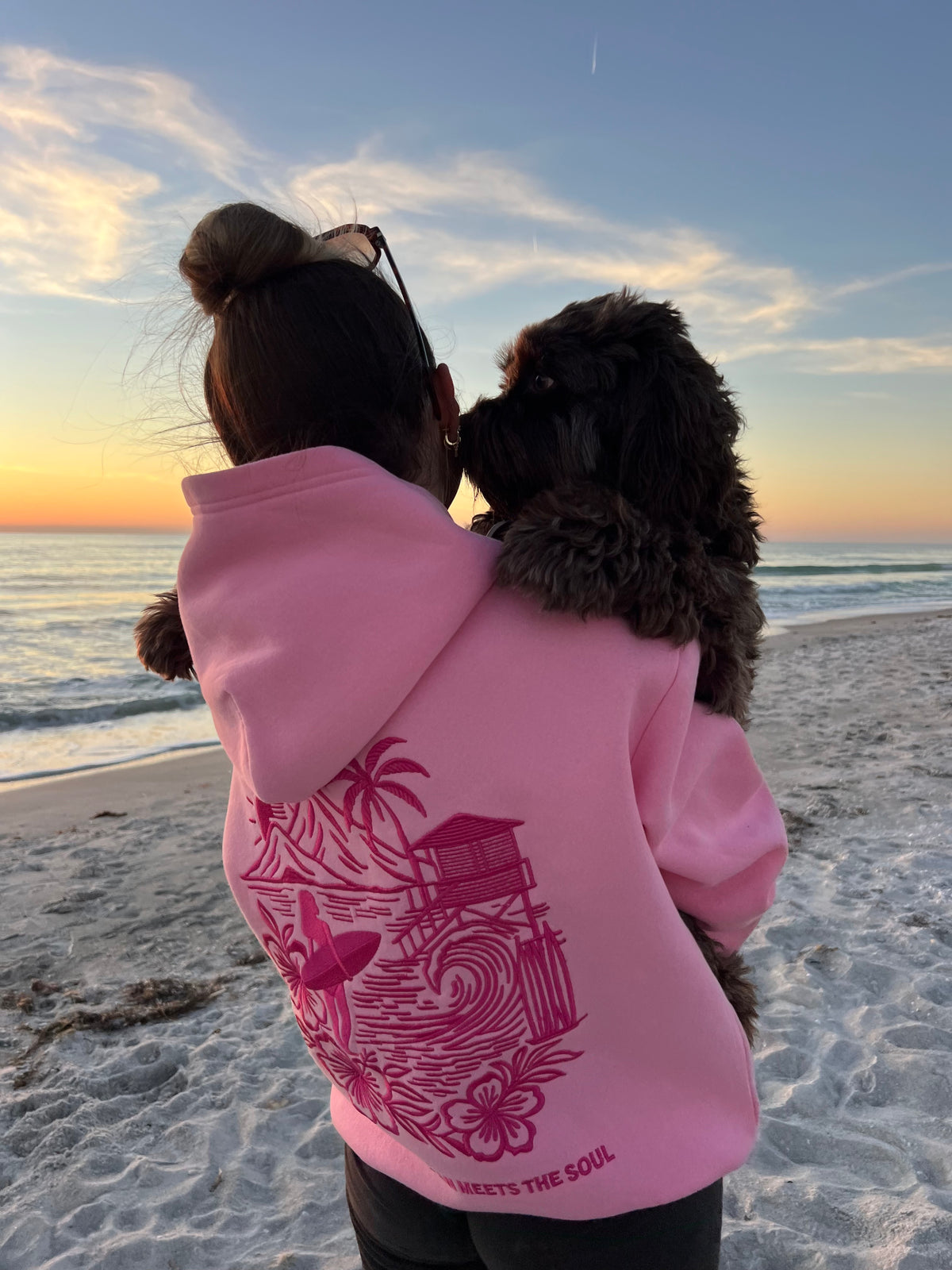 Back of a pink Summit & Coast hoodie with embroidered coastal design on the beach at sunset